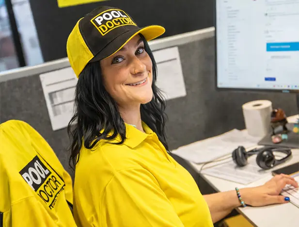A woman wearing a yellow "Pool Doctor" shirt and hat sits at an office desk with a computer, paperwork, and headphones, smiling at the camera.