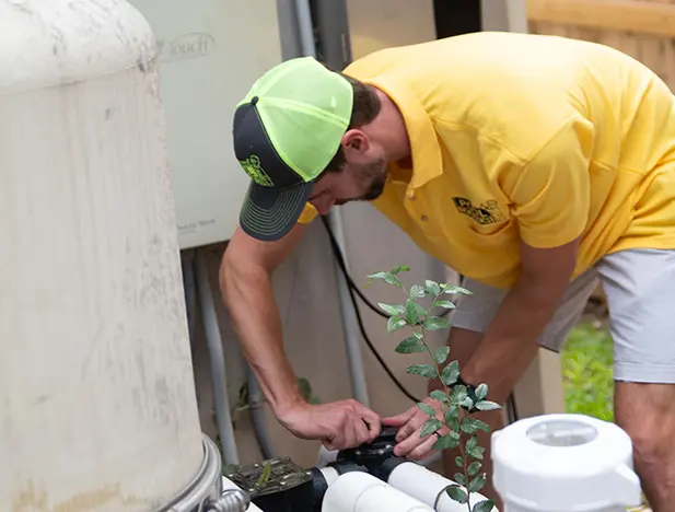 A man wearing a yellow shirt, light shorts, and a neon yellow cap is adjusting equipment outdoors near a large tank. There are plants and pipes visible around him.